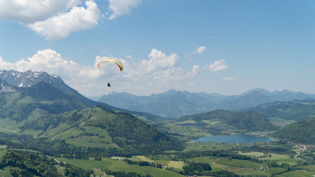 Paragliding in Kössen, Austria with a solo pilot flying above green alpine valleys, lakes, and mountain ridgelines.